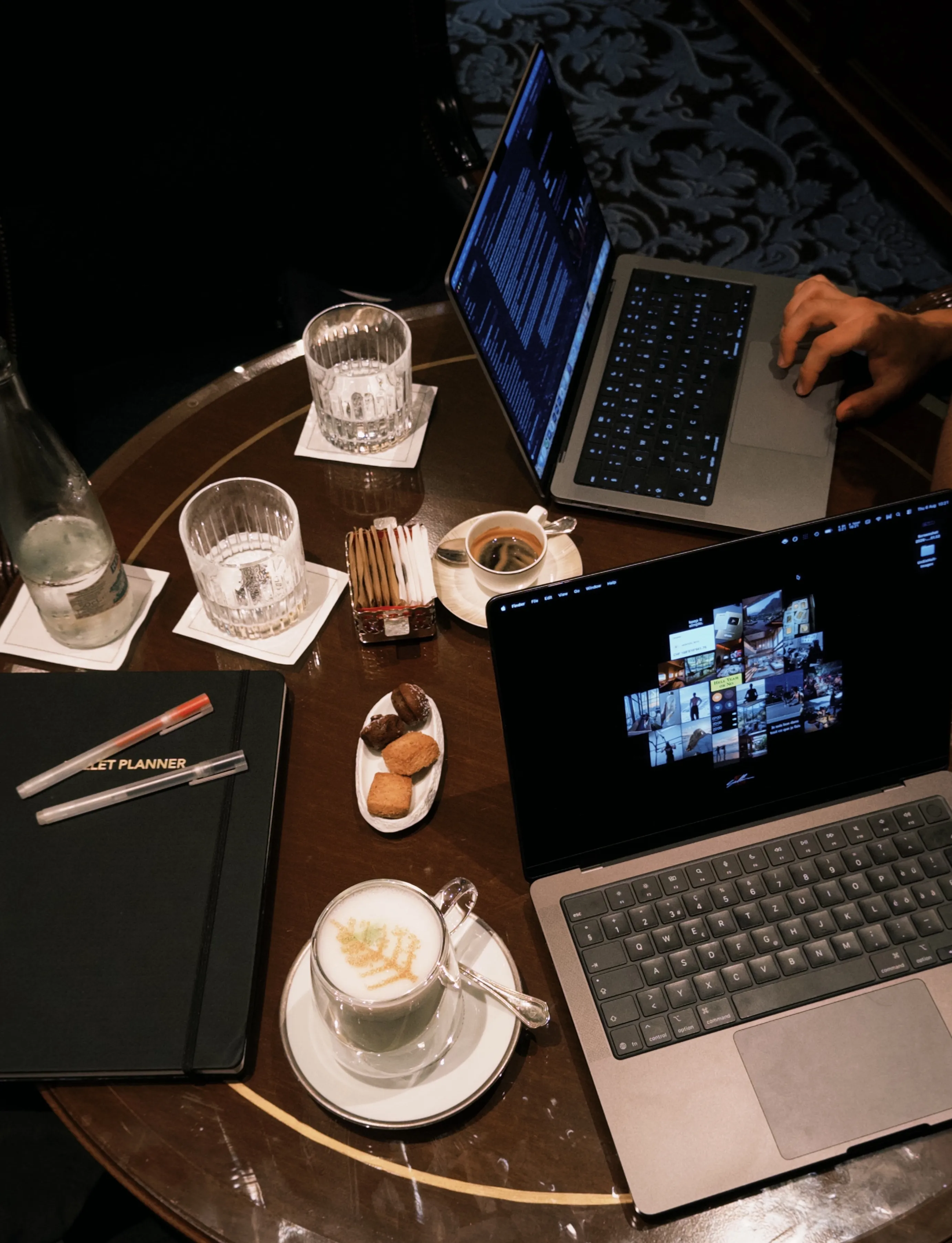 Deux ordinateurs ouverts sur une table de salon, carnet, cafés et carafe d’eau.