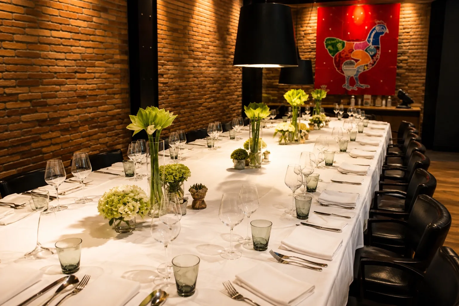Longue table dressée pour un dîner privé, mur de briques et lumière chaude.