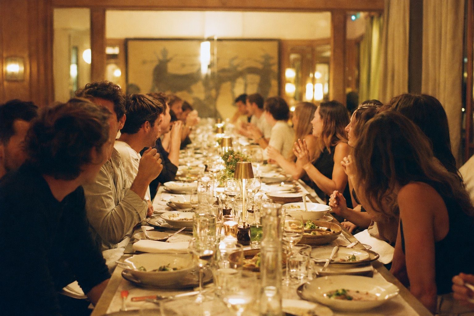 Longue tablée dressée dans une salle boisée à la lumière chaude, convives en conversation de part et d’autre.
